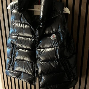 Svart dunväst från Moncler - Säljer min svarta Tibb Down Vest från Moncler med glansig finish och med en klassisk logotyp. Västen har hög krage, två fickor med dragkedja och quiltad design. Stängs med dragkedja framtill.