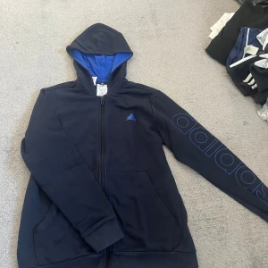 Svart hoodie från Adidas med blå detaljer - Svart hoodie från Adidas med blått foder i huvan och blå Adidas-logga på bröstet. Stora Adidas-tryck längs ena ärmen. Dragkedja framtill och två fickor. 