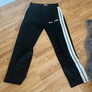 Palm Angels svarta trackpants med ränder - Byxor från Palm Angels med vita dubbla sidoremsor och logga på vänster lår. Byxorna har elastisk midja, raka ben och dragkedja vid benslut. Snygg sportig vibe och passar perfekt till vintern.