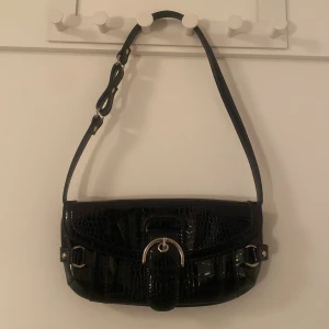Svart axelväska med krokopräglat mönster - Snygg svart axelväska med krokopräglat mönster och silverfärgade detaljer. Axelrem med ett dekorativt spänne framtill. Perfekt storlek för det viktigaste och ger en cool vibe till din outfit.