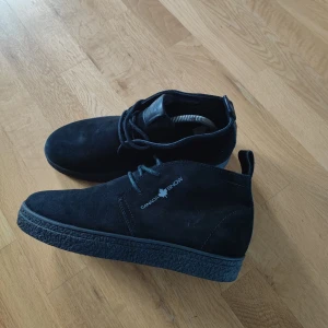 Svarta mockakängor Canada Snow - Snygga svarta kängor från Canada Snow i mjuk mocka med snörning och platt sula. Skorna har en rund tå och diskret logga på sidan. Perfekt för dig som gillar stilrena och klassiska boots med lite streetkänsla.