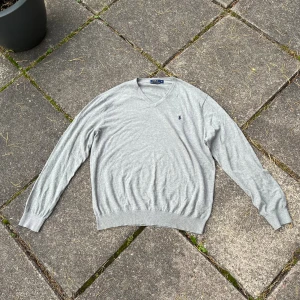 Grå tröja Polo Ralph Lauren - Grå v-ringad tröja från Polo Ralph Lauren i mjuk bomull. Passar mer som en L pga av den passformen. Klassisk design med broderad marinblå logga på bröstet och ribbade muddar vid ärmslut och nederkant. Perfekt för lager-på-lager och chill stil. Bara att skriva om ni har några frågor.