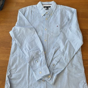 Randig skjorta från Tommy Hilfiger - Snygg blå- och vitrandig skjorta från Tommy Hilfiger med klassisk button down-krage och broderad logga på bröstet. Skjortan har lång ärm, regular fit och är tillverkad i bomull med stretch för extra komfort. Perfekt för en clean och fräsch look.