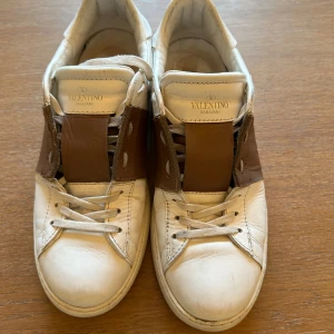 Valentino Garavani vita och bruna sneakers - Snygga sneakers från Valentino Garavani i vitt och brunt skinn. Modellen har rund tå, platt sula och klassisk snörning. Ikonisk design med bred brunt läderpanel över snörningen och subtila detaljer på hälen. Perfekta för dig som gillar exklusiva streetwear-vibbar.