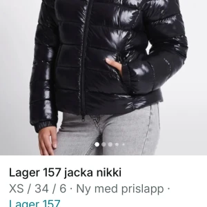 Svart pufferjacka Lager 157 Nikki - Snygg svart pufferjacka från Lager 157, modell Nikki. Jackan har glansig yta, huva och dragkedja framtill. Den är quiltad med breda kanaler och har två sidofickor. Perfekt för kalla dagar och har en modern, loose passform.