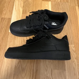 Nike Air Force 1 helsvarta sneakers - Säljer ett par helsvarta Nike Air Force 1 sneakers med chunky snörning och klassisk rund tå. Skorna har en platt sula och är tillverkade i skinn med perforeringar på tån för extra ventilation. Perfekt för dig som gillar stilrena och tidlösa kicks.