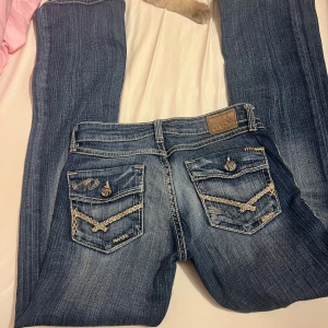 Blå Miss Me jeans med broderade detaljer - Säljer ett par blå Miss Me jeans med snygga slitningar och broderade detaljer på bakfickorna. Jeansen har kontrastsömmar, dekorativa knappar och coola broderier längs fickor och sidor. Passformen är rak och materialet är klassisk jeans med stretch.