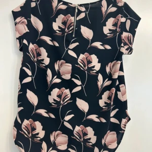 Svart blommig blus med dragkedja - Svart blus med stora rosa och beige blommor, rund halsringning och korta ärmar. Blusen har en synlig dragkedja bak som detalj och är tillverkad i ett mjukt, lätt material. Perfekt för dig som gillar ett romantiskt och trendigt uttryck.