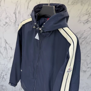 Moncler windbreaker - Snygg mörkblå vindjacka från Moncler med huva och vita ribbade ränder längs axlarna. Jackan har dragkedja framtill, snörning i huvan och Moncler-logga på ärmen. Härligt skick och nästan aldrig använd.