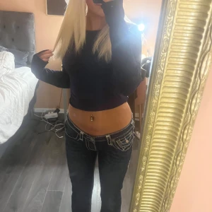 True Religion lågmidjade jeans med kontrastsömmar - Säljer ett par mörkblå True Religion jeans med låg midja och vita kontrastsömmar. Modellen har raka ben men lite utsvängda där nere. Ena knappen är borta på baksidan som ni ser på bilden. Strl 30/s