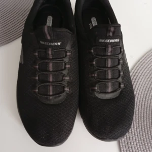 Svarta Skechers sneakers Wide Fit - Svarta sneakers från Skechers med mesh-ovandel och elastisk snörning. Modellen har bred passform och memory foam-innersula för extra komfort. Snyggt diskret S-logga på sidan och platt sula med bra grepp. Perfekt för dig som vill ha stilrena och bekväma skor.