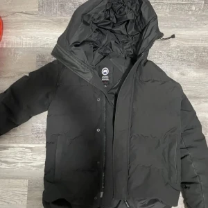 Canada Goose Macmilen  - Svart dunjacka från Canada Goose med huva och klassisk logga på ärmen. Jackan har dragkedja och knappar framtill, två stora fickor och är perfekt för kalla dagar. Snygg och stilren design som håller dig varm hela vintern.