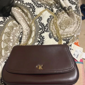 Vinröd axelväska från Calvin Klein - Snygg vinröd axelväska från Calvin Klein i slätt skinn med guldfärgad kedjerem och CK-logga framtill. Väskan har ett rymligt fack och innerficka, perfekt för mobilen och småprylar. Stilren och tidlös design som lyfter vilken outfit som helst. 44% RABATT!! PASSA PÅ KÖP FÖRST!!!