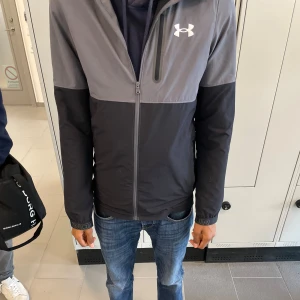 Grå och svart vindjacka Under Armour XS - Snygg vindjacka från Under Armour i grått och svart med huva och hel dragkedja framtill. Jackan har en sportig look, logga på bröstet och detaljer vid ärmsluten.