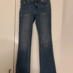 True Religion BECCA Bootcut Jeans - Snygga blå jeans från True Religion, modell BECCA Mid Rise Bootcut. Jeansen har klassisk femficksdesign, markerade sömmar och coola fickdetaljer bak med knappar. Bootcut-modell som ger en schysst siluett och passar perfekt till sneakers eller boots. Storlek: 27