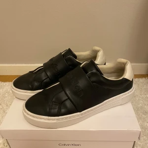 Svarta sneakers från Calvin Klein - Snygga svarta sneakers från Calvin Klein med breda remmar över foten där loggan är präglad. 
