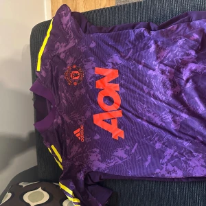 Manchester United lila målvaktströja Adidas - Säljer en lila Manchester United målvaktströja från Adidas med orangea detaljer och klubbmärke på bröstet. Tröjan har korta ärmar med gula ränder på axlarna och ett unikt mönster över hela tyget. Perfekt för dig som vill sticka ut på planen.