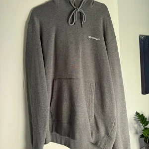 Grå Off-White hoodie med tryck - Säljer en grå hoodie från Off-White med vit logga på bröstet och stort grafiskt tryck på ryggen. Tröjan har huva med snörning, känguruficka och är tillverkad i mjuk bomull. Perfekt för dig som gillar streetwear och vill sticka ut. Köpt för 4500, prisförslag uppskattas!!