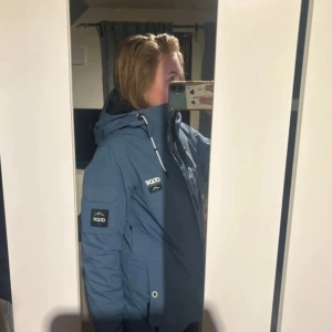 Blå skidjacka från Dope med huva - Säljer en blå skidjacka från Dope. Jackan har huva med snörning, flera fickor med dragkedja och coola metallöglor. Perfekt för dig som gillar streetwear och outdoor-vibe. Använd enbart två gånger så i  bra skick, den är mellanfodrad.