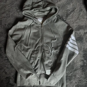 Grå hoodie från TB Ton Bdoune - Snygg grå hoodie från Thom browne med dragkedja och huva. Tröjan har vita ränder på ena ärmen, snörning vid huvan och en vit patch med text på framsidan. Perfekt för chill dagar och streetwear-vibe. Ända felet med tröjan är att snöret är lite skumt som man ser på bilden!