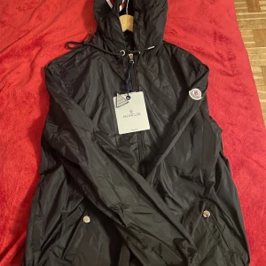 Svart vindjacka från Moncler - Pris kan diskuteras!! Svart vindjacka från Moncler med huva och snygga detaljer som snörning och dragkedja framtill. Jackan har Moncler-logga på ärmen och silverfärgade knappar vid fickorna. Materialet är lätt och syntetiskt, perfekt för blåsiga dagar. Du kan scanna loggan på kanten av armen. 