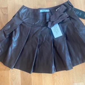 Brun kortkjol i skinn från NA-KD - Supersnygg brun kortkjol i skinnimitation från NA-KD x Sofia Coelho. Kjolen har ett medföljande bälte, bred linning och snygga veck som ger extra volym. Perfekt för dig som vill ha en trendig och edgy look med coola detaljer. HELT NY MED PÅSE OCH TAGGAR (har två därför säljer jag ena), för fler bilder passa på fråga