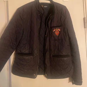 Morris quiltad jacka marinblå M - Quiltad marinblå jacka från Morris med broderad detalj på bröstet. Jackan har ståkrage, dragkedja och knappar framtill samt svarta detaljer vid fickorna. Perfekt för kyligare dagar och ger en klassisk look.