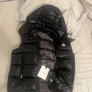 Svart dunväst från Moncler med huva - Svart, glansig dunväst från Moncler med avtagbar huva och två dragkedjeförsedda fickor. Västen har quiltad design och klassisk Moncler-logga på bröstet. Insidan har en cool serietidningsetikett och Moncler-märke. Perfekt för att maxa din streetstyle.