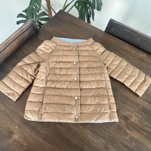 Reversibel pufferjacka från Herno - Snygg reversibel pufferjacka från Herno med raka, quiltade paneler och guldfärgade knappar. Ena sidan är ljusblå och andra sidan är beige, vilket gör den superenkel att matcha. Jackan har trekvartsärmar och en stilren, kraglös design. Perfekt för dig som gillar att variera din look.