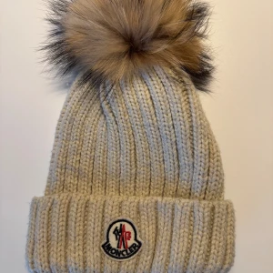 Beige Moncler mössa med pälsboll - Ribbstickad beige mössa från Moncler med stor, fluffig avtagbar pälsboll i bruna och svarta toner. Mössan har uppvikt kant och klassisk Moncler-logga framtill. Perfekt accessoar för kalla dagar med exklusiv känsla.