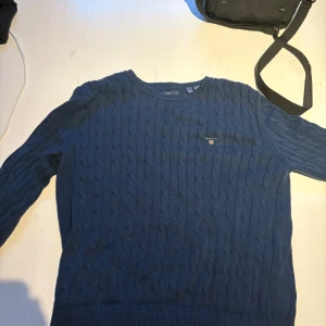 Blå stickad tröja från GANT - Säljer en snygg blå stickad tröja från GANT. Tröjan har ett klassiskt flätmönster och rund halsringning. Bra skick använd då gånger! 
