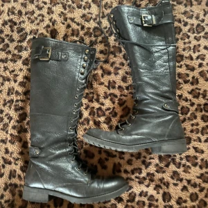 Svarta höga kängor i skinn - Säljer ett par svarta höga kängor i skinn med snörning hela vägen upp och spänne upptill. De har platt sula och rund tå, perfekt för en edgy look. Passar till både jeans och kjol för en cool stil.