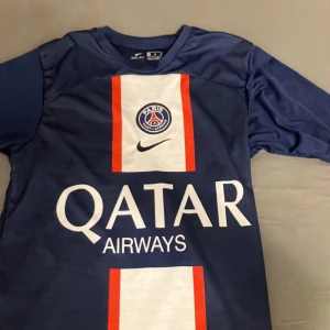 PSG Mbappé matchtröja Nike blå - Snygg Paris Saint-Germain fotbollströja med Mbappé och nummer 7 på ryggen. Tröjan är marinblå med vita och röda detaljer, PSG-emblem på bröstet och Qatar Airways som sponsor. Tillverkad i lätt och ventilerande polyester, perfekt för fotbollsplanen.