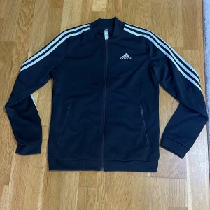 Svart Adidas track jacket  - Svart Adidas track jacket med klassiska vita ränder längs ärmarna och broderad logga på bröstet. Jackan har ribbad krage, muddar och nederkant samt två fickor med dragkedja. Materialet är mjuk polyester och modellen är normal i passningen.