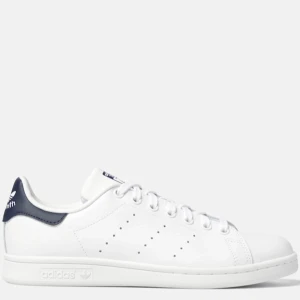 Adidas Stan Smith vita sneakers - Klassiska Adidas Stan Smith sneakers i vitt skinn med marinblå detalj på hälen. Snygg minimalistisk design med perforerade ränder på sidorna och rund tå. Skosnören ser gråare ut på bilderna