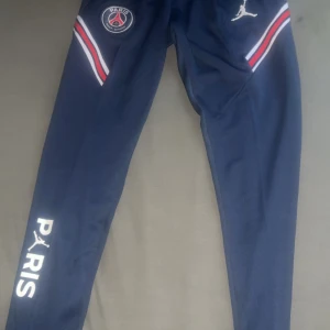 PSG x Jordan mörkblå träningsbyxor M - Snygga mörkblå träningsbyxor från Jordan x PSG med vita och röda detaljer på benen, PSG-logga och Jordan-logga. Byxorna har elastisk midja, smal passform och PARIS-tryck längs benet. Perfekta för dig som gillar sportig stil.