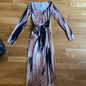 Långklänning med abstrakt mönster och knyt - Långklänning i beige/rosa och svart med abstrakt mönster. Klänningen har lång ärm, rund halsringning och ett snyggt knyt i midjan. Materialet är mjukt och följsamt, perfekt för dig som gillar en unik look med coola detaljer. Använd en gång! 