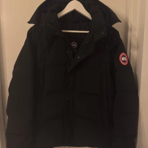 Canada Goose Wyndham  - Svart pufferjacka från Canada Goose med ikonisk patch på ärmen. Jackan har huva, dragkedja och tryckknappar framtill. Fylld med dun och fjädrar för extra värme, perfekt för kalla dagar. Klassisk, stilren design med normal passform och långa ärmar.