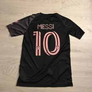 Inter Miami Messi matchtröja Adidas S - Snygg svart och mörkgrå Inter Miami fotbollströja från Adidas med rosa detaljer, Messi och nummer 10 på ryggen. Tröjan har korta ärmar, klubbmärke och sponsorloggor framtill. Tillverkad i lätt och ventilerande AEROREADY-material.