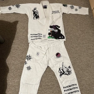 Vit grappling gi med tryck, storlek 0w=a1 (inte min bild) - Säljer en vit grappling gi från Shoyoroll med unika tryck och illustrationer i svart och lila. Jackan har texten 'PANCRATTY!' och brottningsmotiv, samt snörning i midjan på byxorna. Perfekt för dig som tränar grappling eller MMA.