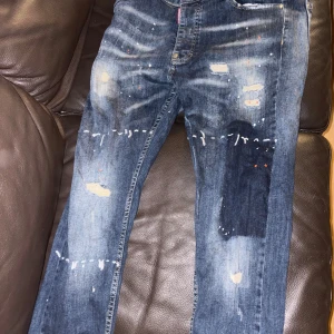 Dsquared2 jeans med färgstänk - Säljer ett par blå jeans från Dsquared2 med coola färgstänk och slitningar. Jeansen har klassisk femficksdesign, knappgylf och orange kontrastsömmar. Materialet är robust denim och passformen är rak med normal midja. Perfekt för dig som gillar streetstyle.