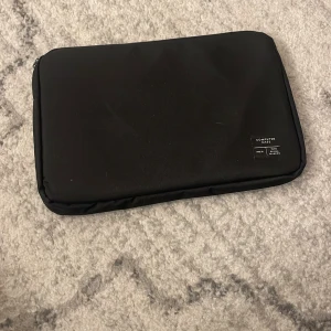 Svart datorväska från WISIO MAKERS - En stilren svart datorväska från WISIO MAKERS med minimalistisk design. Väskan har en mjuk, vadderad utsida och dragkedja längs kanten. Perfekt för att skydda din laptop på språng.