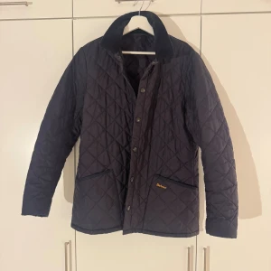 Barbour Liddesdale navy - Stilren svart quiltad jacka från Barbour med klassisk krage i manchester och tryckknappar framtill. Jackan har två fickor med Barbour-logga och ett tidlöst rutmönster i quiltningen. Perfekt för dig som vill ha en snygg och funktionell jacka.