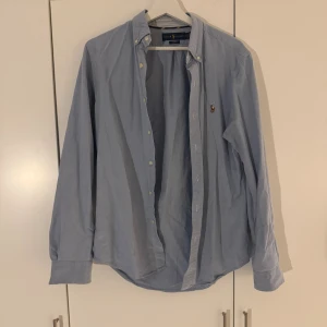 Oxfordskjorta Ralph Lauren Slim fit - Klassisk ljusblå skjorta från Ralph Lauren med broderad logga på bröstet. Skjortan har lång ärm, knappar framtill och en snygg krage. Perfekt för dig som gillar stilrena och tidlösa plagg med en touch av preppy.