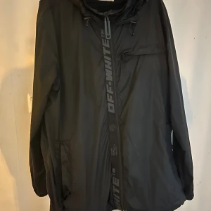 Svart vindjacka från Off-White - Svart vindjacka från Off-White med huva och coola broderade detaljer på ryggen. Jackan har dragkedja framtill med Off-White-tryck, snörning i nederkant och flera fickor. Materialet är lätt och syntetiskt, perfekt för blåsiga dagar. Snygg streetstil med loose passform. Lite trasig vid ena fickan och ett litet hål se bilder