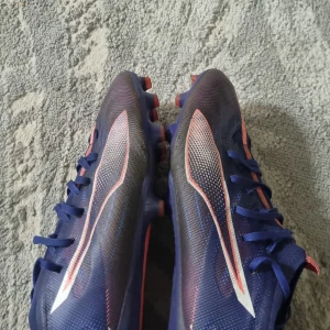 Blå fotbollsskor med rosa  detaljer - Säljer ett par snygga blå fotbollsskor från Puma med  råsa och vita detaljer. Skorna har snörning, platt sula med fasta dobbar och är tillverkade i syntetmaterial för bästa grepp på planen. Perfekta för dig som vill sticka ut på matchen!