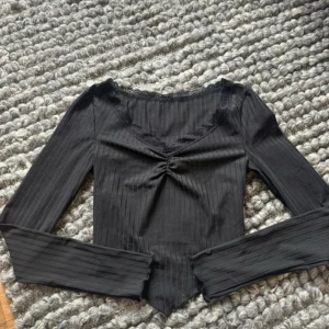 Svart ribbad långärmad topp med spets - Svart långärmad topp med ribbat tyg och v-ringning dekorerad med spetskant. Toppen har en snygg rynkdetalj framtill och en något spetsig nederkant. Perfekt för dig som gillar stilrena och feminina detaljer.