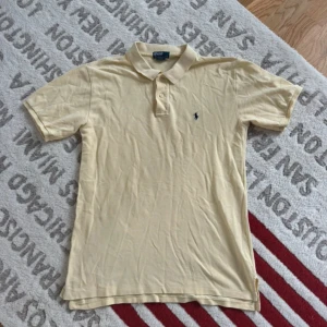 Gul pikétröja från Polo Ralph Lauren - Klassisk gul pikétröja från Polo Ralph Lauren med korta ärmar och broderad logga på bröstet. Tröjan har krage och två knappar framtill. Tillverkad i bomull för en skön och luftig känsla. Perfekt för en clean och stilren look.