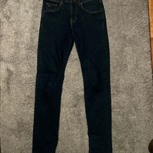 Mörkblå jeans från J.Lindeberg 29/32 - Snygga mörkblå jeans från J.Lindeberg med klassisk femficksdesign och diskret JL-logga på myntfickan. Jeansen har smal passform och raka ben, tillverkade i mjukt denim med kontrastsömmar. Perfekta för dig som gillar stilrena och moderna jeans.