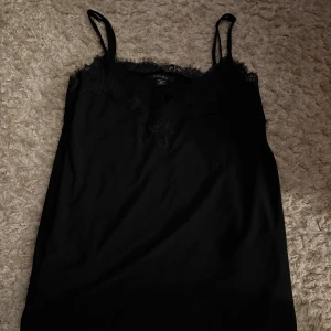 Svart linnetopp med spets från Amisu - Säljer en svart linnetopp från Amisu med tunna axelband och v-ringning dekorerad med snygg spets. Toppen har en stilren och enkel design som känns både trendig och klassisk. Perfekt att styla med jeans eller kjol för en cool look.